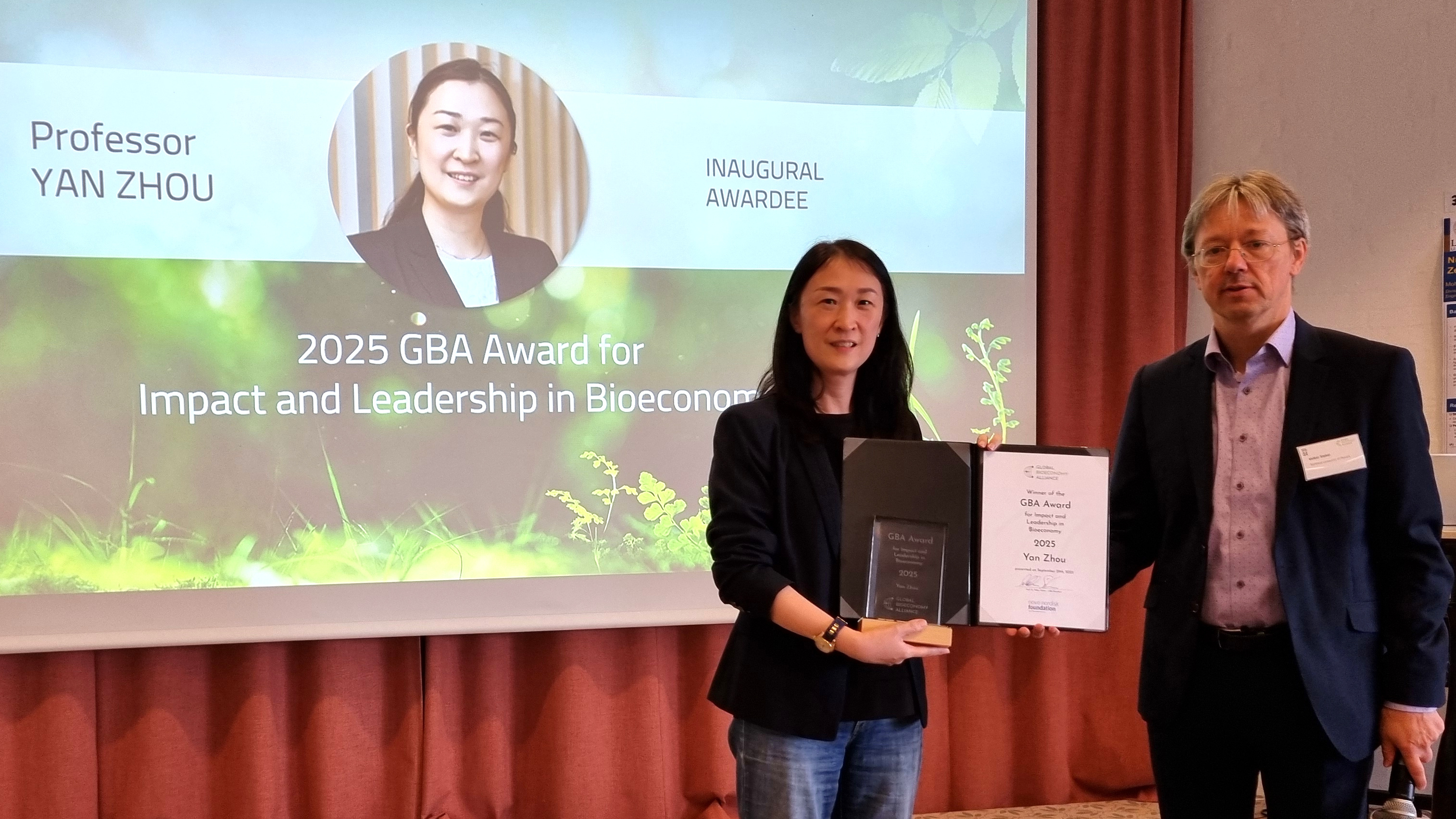 A man and a woman are standing in front of a projetion screen, the slide saying "Professor Yan Zhou, Inaugural Awardee". The woman is in the center of the image, presenting her award certificate and the trophy.